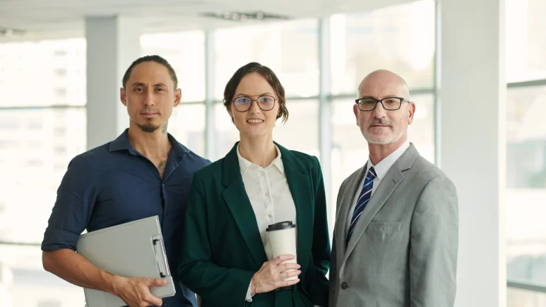 Three business professionals standing together in a bright, modern office. On the left, a man in a dark blue shirt holds a gray clipboard; in the center, a woman wearing glasses and a green blazer holds a coffee cup; and on the right, a bald man with glasses wears a gray suit and tie.