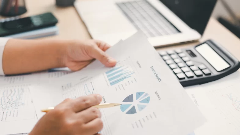 A close-up of a person's hands holding a document titled "Finance Report" containing bar graphs and a pie chart. The person is holding a gold pen, and a calculator and laptop are visible on the wooden desk in the background.