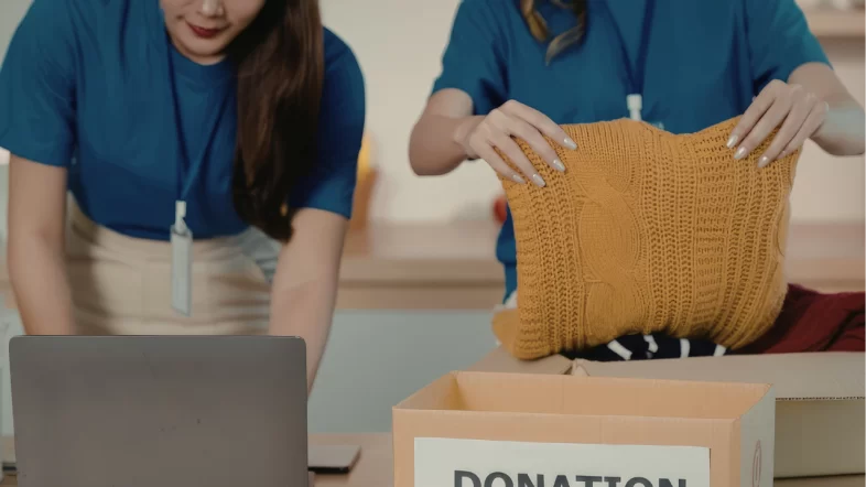 A close-up view of a person folding a yellow knit sweater to place into a cardboard box labeled "DONATION." Another person is partially visible on the left, working on a laptop.