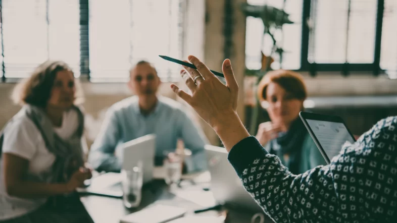 A close-up focus on a person's hand holding a pen and gesturing in the air during a discussion. In the blurred background, other colleagues are seated at a table, listening.