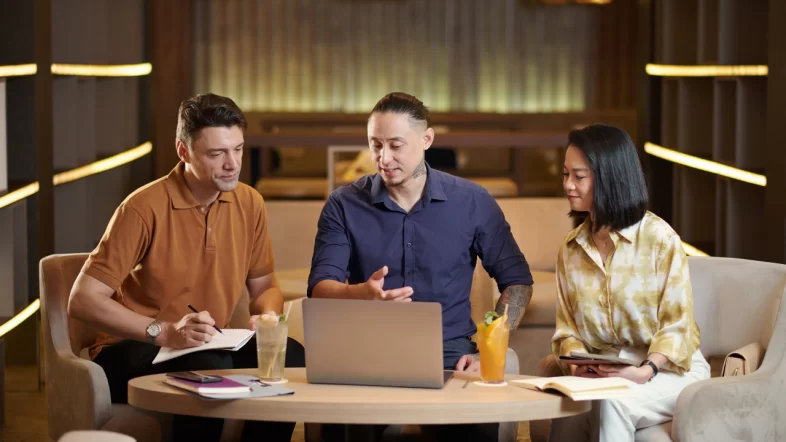 Three colleagues having a casual meeting at a low round table. A man in a dark blue shirt gestures towards a laptop screen, while a man in an orange polo shirt and a woman look on attentively. Drinks and notebooks are present on the table.