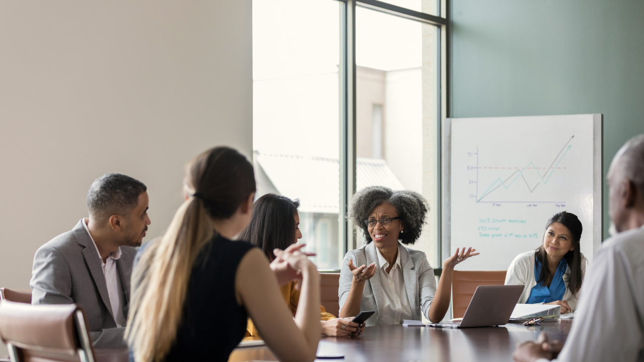 A diverse group of people seated at a conference table during a meeting. A woman with gray curly hair and glasses is speaking and gesturing with open hands to her colleagues across the table, with a whiteboard visible in the background.