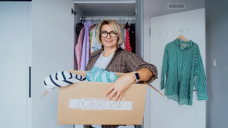 A smiling woman with blonde hair and glasses stands in front of an open closet, holding a cardboard box labeled "DONATION" that is filled with folded clothes.